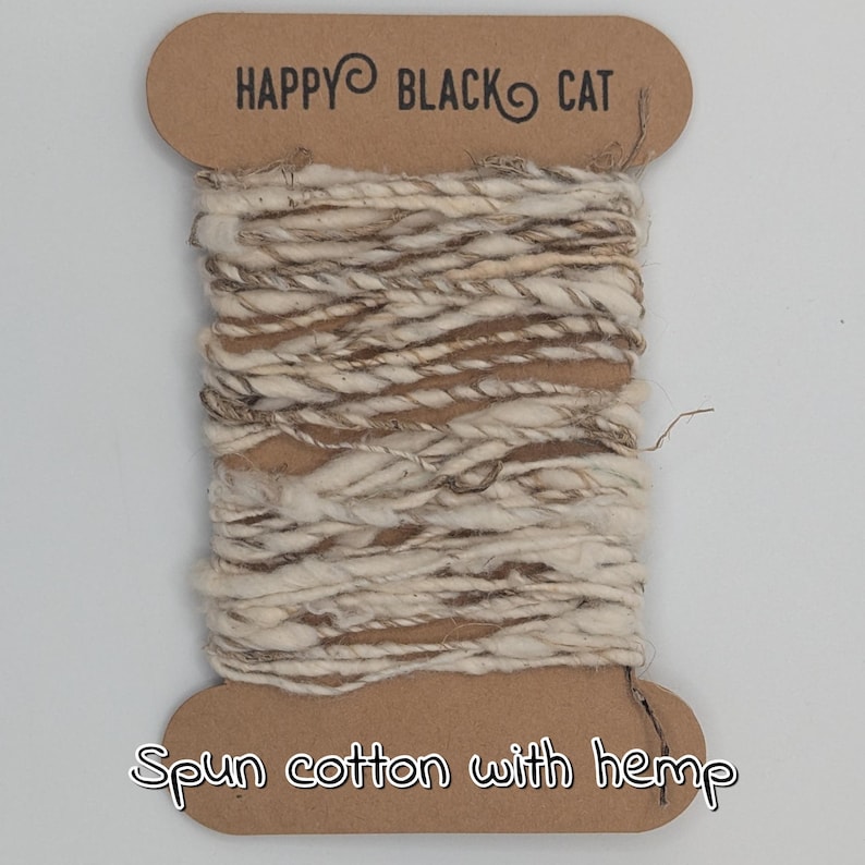 Peut inclure: Une bobine de fil de coton fil&eacute; avec des fibres de chanvre. Le fil est un m&eacute;lange de tons cr&egrave;me et marron, enroul&eacute; autour d'une bobine en carton. Le texte "HAPPY BLACK CAT" est imprim&eacute; en haut de la bobine, et "Spun cotton with hemp" en bas.