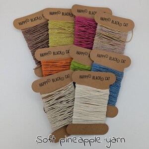 May include: A collection of colorful yarn spools. Each spool is labeled "HAPPY BLACK CAT" and holds yarn in shades of brown, yellow, pink, tan, orange, green, blue, and white. The text "Soft pineapple yarn" is at the bottom.