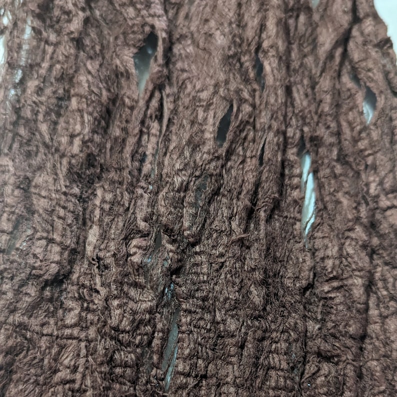 May include: Close-up of a brown, textured fabric with a rough, shredded appearance. The fabric has several holes and a crinkled texture, suggesting a distressed or aged aesthetic. The material appears to be a decorative element.