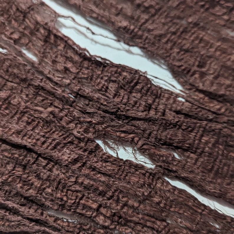 May include: Close-up of a textured, dark brown fabric with a distressed appearance. The fabric has a woven pattern with visible frayed edges and several irregular openings, creating a worn, vintage aesthetic. The background is a bright white.