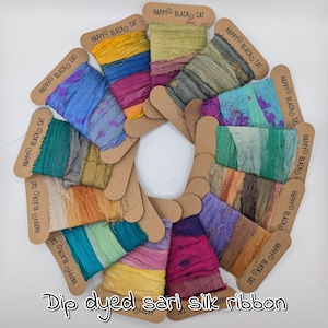 May include: A selection of colourful dip-dyed sari silk ribbon spools, each on a cardboard tag with "HAPPY BLACK CAT" printed on it. The ribbons display a range of hues, including blue, green, and purple. The text "Dip dyed sari silk ribbon" is at the bottom.