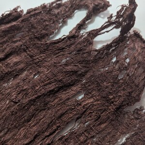 May include: Close-up of a piece of dark brown fabric with a distressed, textured appearance. The fabric has a crinkled, shredded design with visible holes and frayed edges, creating a rustic aesthetic.