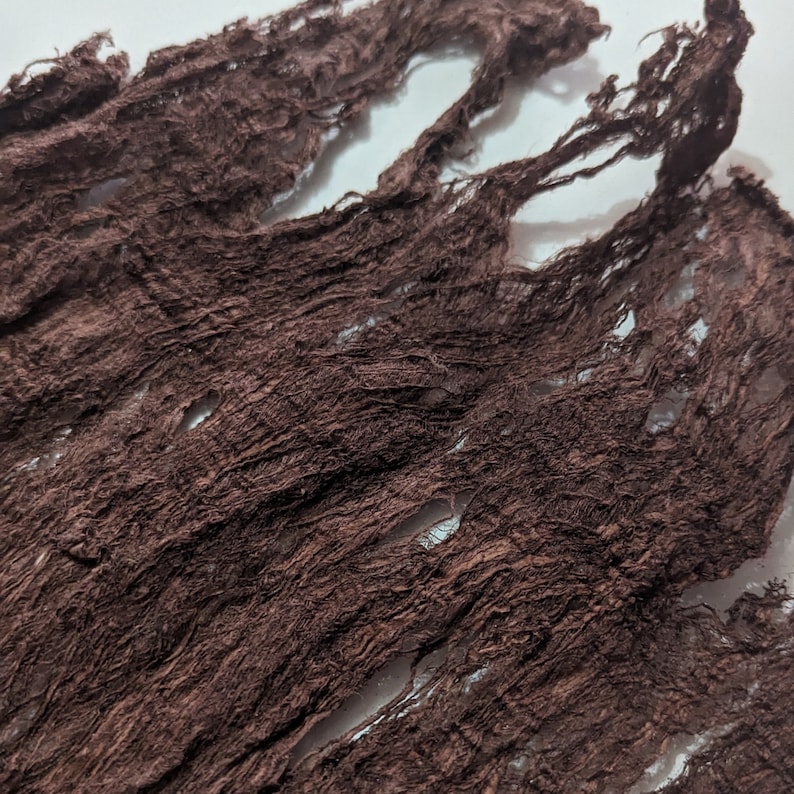 May include: Close-up of a piece of dark brown fabric with a distressed, textured appearance. The fabric has a crinkled, shredded design with visible holes and frayed edges, creating a rustic aesthetic.