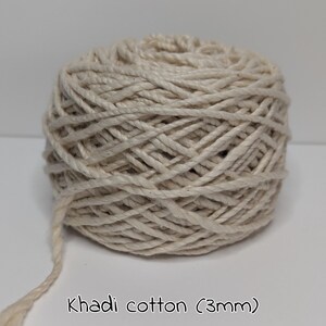 Peut inclure: Une pelote de fil de coton Khadi blanc cass&eacute;, de 3 mm d'&eacute;paisseur, est pr&eacute;sent&eacute;e sur un fond uni. Le fil est bien enroul&eacute;, avec un brin l&acirc;che qui s'&eacute;tend de la pelote. Le texte "Khadi cotton (3mm)" est visible en bas.