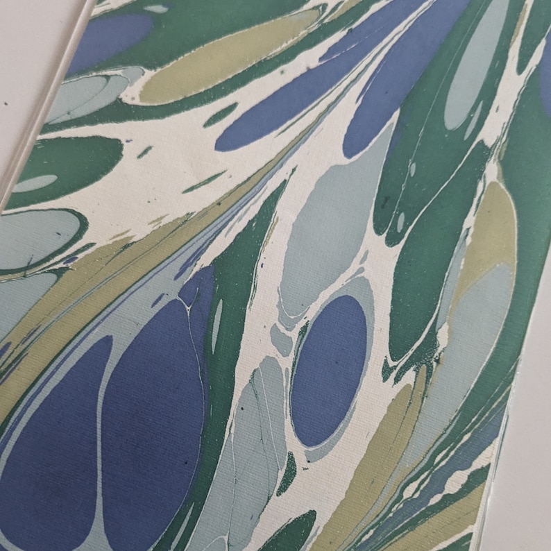 May include: Close-up of marbled paper with a flowing pattern. The design features organic shapes in shades of blue, green, and beige against a white background. The paper has a textured appearance, with visible swirls and veins.