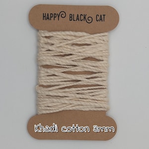 Peut inclure: Une bobine de corde en coton Khadi de couleur naturelle, de 3 mm d'&eacute;paisseur, est pr&eacute;sent&eacute;e sur un fond blanc. La bobine est &eacute;tiquet&eacute;e avec le texte "HAPPY BLACK CAT" en haut et "Khadi cotton 3mm" en bas. La corde est bien enroul&eacute;e.