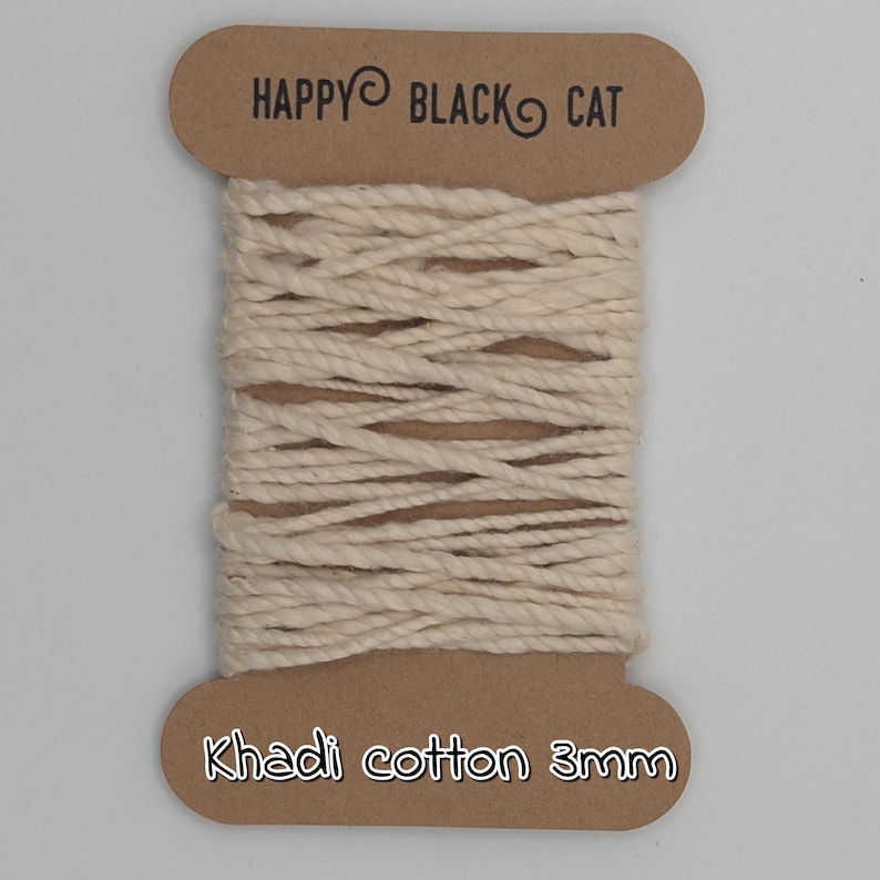 Peut inclure: Une bobine de corde en coton Khadi de couleur naturelle, de 3 mm d'&eacute;paisseur, est pr&eacute;sent&eacute;e sur un fond blanc. La bobine est &eacute;tiquet&eacute;e avec le texte "HAPPY BLACK CAT" en haut et "Khadi cotton 3mm" en bas. La corde est bien enroul&eacute;e.