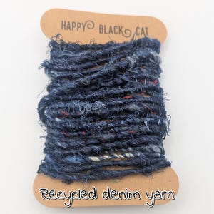 May include: A spool of recycled denim yarn in shades of blue, with flecks of red and white. The yarn is wound around a cardboard spool with the text "HAPPY BLACK CAT" and "Recycled denim yarn".