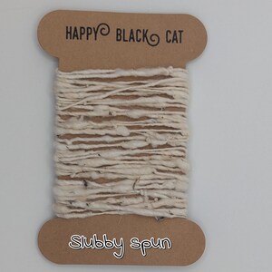 Peut inclure: Une bobine de fil blanc cass&eacute;, fil&eacute; grossi&egrave;rement. Le fil est enroul&eacute; autour d'une &eacute;tiquette en carton avec le texte "HAPPY BLACK CAT" en haut et "Stubby spun" en bas. Le fil a un aspect textur&eacute; et l&eacute;g&egrave;rement irr&eacute;gulier.