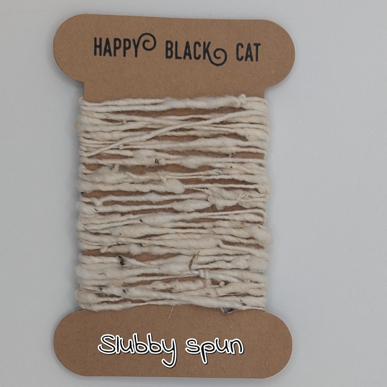 Peut inclure: Une bobine de fil blanc cass&eacute;, fil&eacute; grossi&egrave;rement. Le fil est enroul&eacute; autour d'une &eacute;tiquette en carton avec le texte "HAPPY BLACK CAT" en haut et "Stubby spun" en bas. Le fil a un aspect textur&eacute; et l&eacute;g&egrave;rement irr&eacute;gulier.
