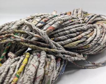 May include: Close-up of a ball of yarn made from rolled newspaper strips. The yarn is a mix of gray, white, and colorful newsprint, with visible text and images. The yarn is tightly wound, creating a textured, organic shape.