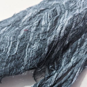 May include: Close-up of a textured, dark gray fabric with a frayed, layered appearance. The fabric has a rough, uneven surface with visible fibers and a few small red threads. The material appears to be soft and pliable.