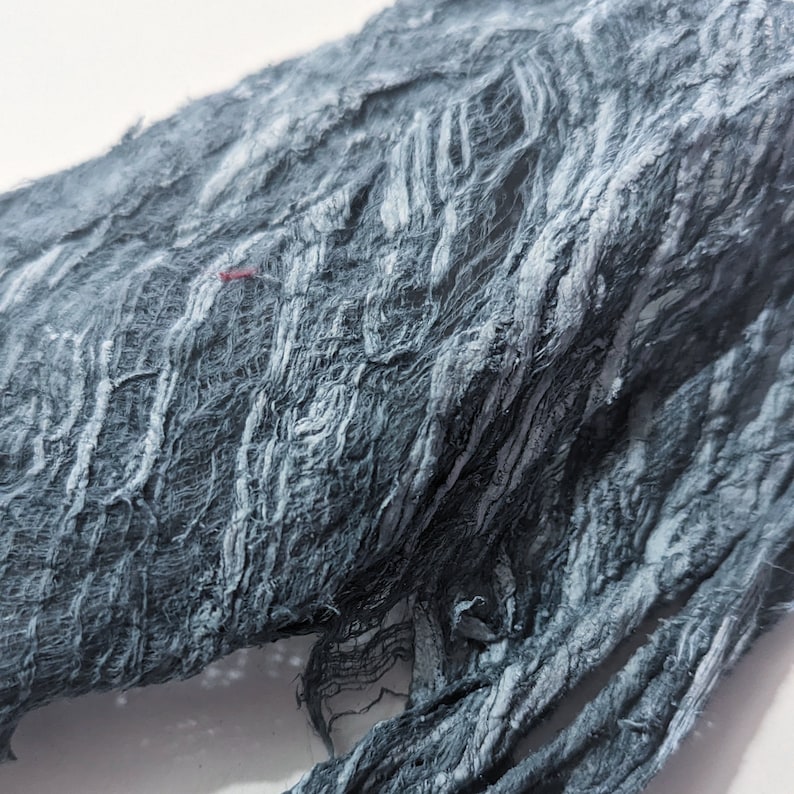 May include: Close-up of a textured, dark gray fabric with a frayed, layered appearance. The fabric has a rough, uneven surface with visible fibers and a few small red threads. The material appears to be soft and pliable.