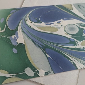 May include: A close-up of a rectangular piece of marbled paper with a swirling pattern. The design features shades of blue, green, and white, creating an abstract, organic look. The paper has a textured surface, suitable for stationery or art projects.