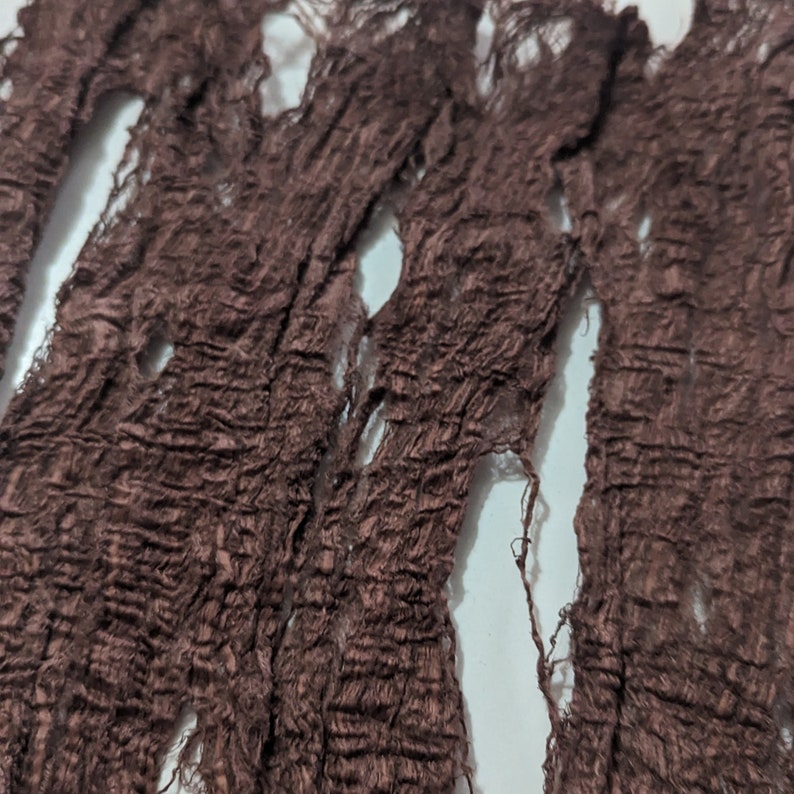 May include: Close-up of a dark brown, shredded fabric with a textured, wrinkled appearance. The fabric has several holes and frayed edges, creating a distressed look. The material appears to be lightweight and possibly used for decorative purposes.