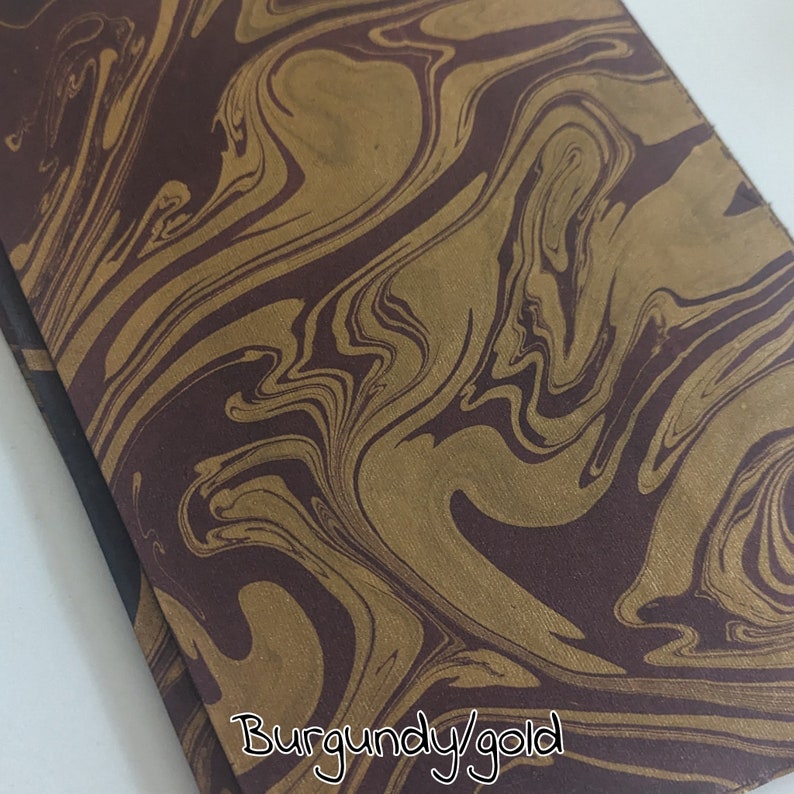 May include: Close-up of a burgundy and gold marbled paper. The swirling pattern features a rich burgundy base with flowing gold accents. The text "Burgundy/gold" is written in a script font at the bottom of the image.