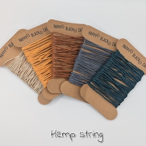 Puede incluir: Cinco carretes de hilo de cáñamo en varios colores: natural, naranja, marrón, gris azulado y verde azulado. Cada carrete está montado en una tarjeta marrón con el texto "HAPPY BLACK CAT". Debajo de los carretes está escrito "Hemp string".