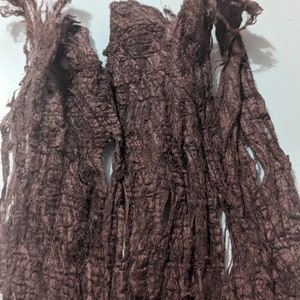 May include: Close-up of a dark brown, textured fabric with a frayed, shredded appearance. The material has a rough, uneven surface with multiple openings and a slightly shiny finish. The fabric appears to be a scarf or shawl.