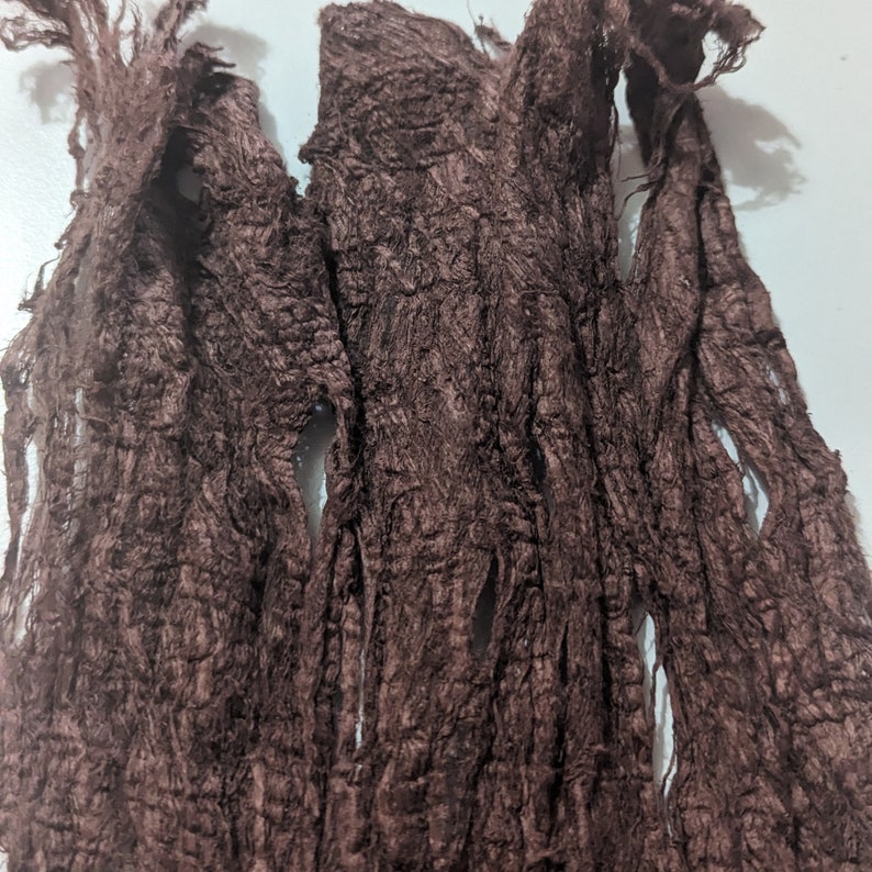 May include: Close-up of a dark brown, textured fabric with a frayed, shredded appearance. The material has a rough, uneven surface with multiple openings and a slightly shiny finish. The fabric appears to be a scarf or shawl.