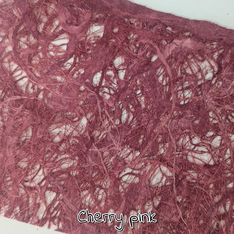 May include: Close-up of a sheet of cherry pink handmade paper. The paper has a textured, fibrous appearance with a network of interwoven strands. The text "Cherry pink" is written in a cursive font at the bottom.