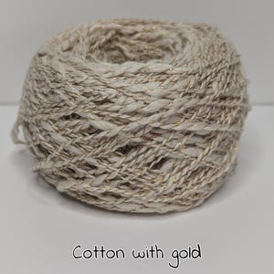 Peut inclure: Pelote de fil de coton beige avec des fils m&eacute;talliques dor&eacute;s. Le fil est &eacute;troitement enroul&eacute;, pr&eacute;sentant une surface textur&eacute;e. Le texte "Cotton with gold" est au bas de l'image.