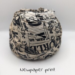 Peut inclure: Une boule de tissu avec un imprim&eacute; journal. Le tissu est de couleur cr&egrave;me avec du texte noir et les bords sont effiloch&eacute;s. Les mots "WORLD" et "team, an" sont visibles. Le texte "Newpaper print" est &eacute;crit en dessous.