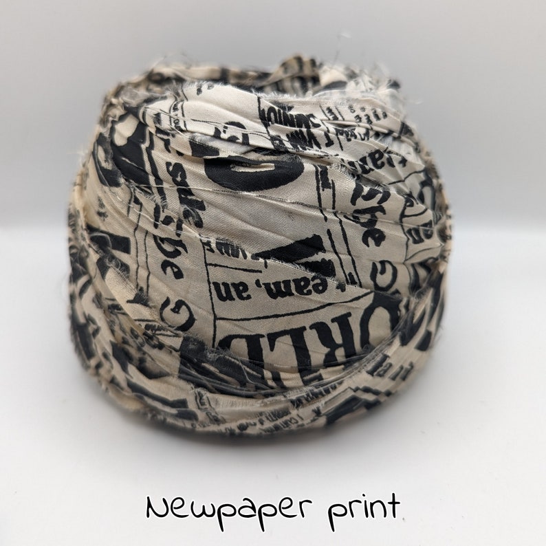 Peut inclure: Une boule de tissu avec un imprim&eacute; journal. Le tissu est de couleur cr&egrave;me avec du texte noir et les bords sont effiloch&eacute;s. Les mots "WORLD" et "team, an" sont visibles. Le texte "Newpaper print" est &eacute;crit en dessous.