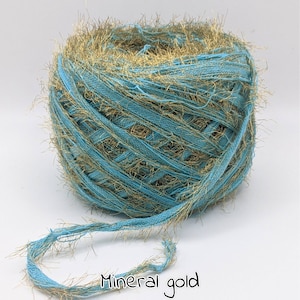 May include: A ball of teal and gold yarn. The yarn has a textured appearance with gold fringe. The text "Mineral gold" is written in a handwritten font at the bottom of the image. This yarn is suitable for various crafting projects, such as knitting or crochet.