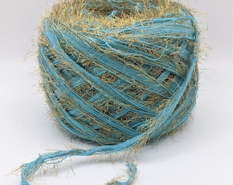 May include: A ball of teal and gold yarn. The yarn has a textured appearance with gold fringe. The text "Mineral gold" is written in a handwritten font at the bottom of the image. This yarn is suitable for various crafting projects, such as knitting or crochet.