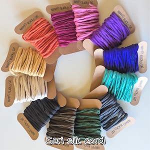 May include: A collection of eleven spools of sari silk cord in various colours including orange, magenta, pink, purple, blue, teal, dark grey, green, brown, black, and cream. Each spool is made of brown cardboard labelled Happy Black Cat.