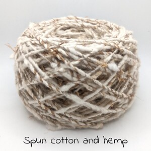 Peut inclure: Une pelote de fil de coton et de chanvre fil&eacute;. Le fil est un m&eacute;lange de fibres blanches et beiges, cr&eacute;ant un aspect textur&eacute; et naturel. Le texte "Spun cotton and hemp" est &eacute;crit en bas de l'image.