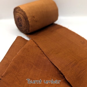 May include: A roll of burnt umber-colored fabric sits next to a piece of the same fabric, unrolled. The fabric has a slightly textured appearance. The words "Burnt umber" are written in a cursive font at the bottom of the image.