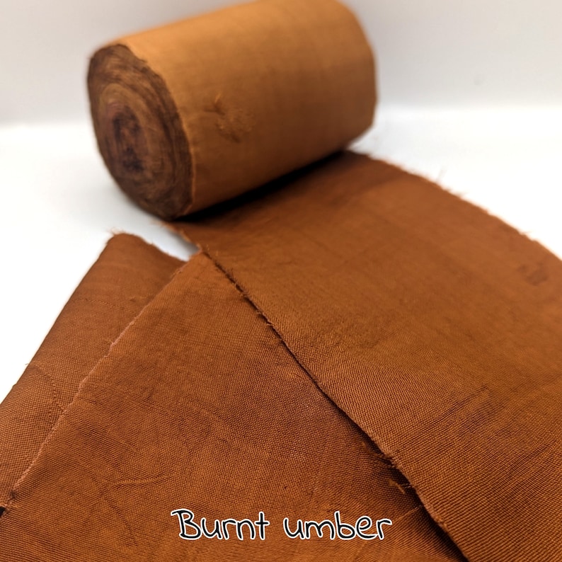 May include: A roll of burnt umber-colored fabric sits next to a piece of the same fabric, unrolled. The fabric has a slightly textured appearance. The words "Burnt umber" are written in a cursive font at the bottom of the image.