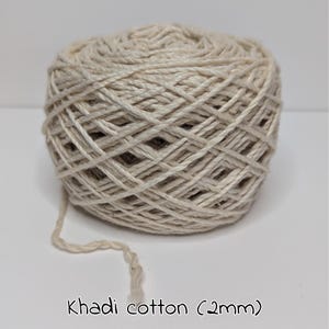 Peut inclure: Pelote de fil de coton Khadi, d'une &eacute;paisseur de 2 mm, de couleur beige naturel. Le fil est bien enroul&eacute;, avec un brin l&acirc;che qui s'&eacute;tend de la pelote. Le texte "Khadi cotton (2mm)" est &eacute;crit en dessous.