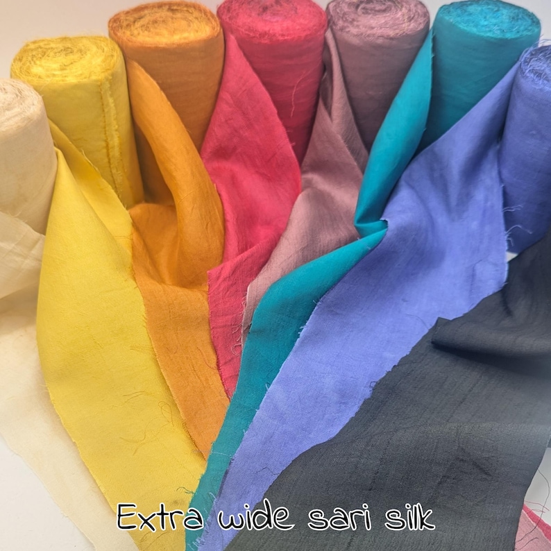 May include: A collection of extra wide sari silk fabric in various colors, including yellow, orange, pink, teal, purple, and gray. The fabric is rolled up and displayed with the edges fanned out, showcasing the different hues. The text "Extra wide sari silk" is visible.