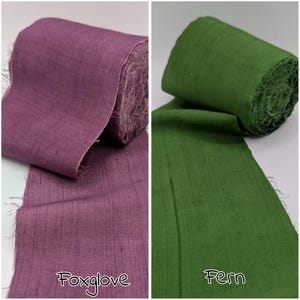 May include: Two rolls of fabric, one in a deep purple color labeled "Foxglove" and the other in a green color labeled "Fern". The fabric appears to be a woven material with a slightly textured surface. The edges of the fabric are frayed.