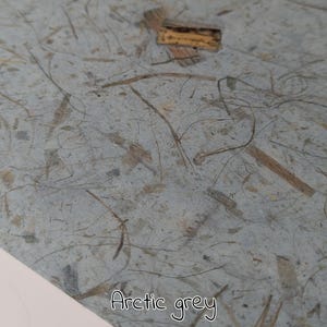 May include: Close-up of arctic grey paper with embedded natural fibers and small wood pieces. The paper has a textured surface and the words "Arctic grey" are written in a cursive font at the bottom.