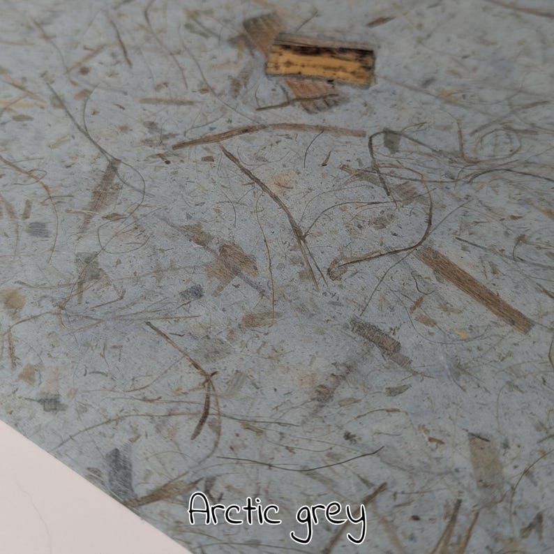 May include: Close-up of arctic grey paper with embedded natural fibers and small wood pieces. The paper has a textured surface and the words "Arctic grey" are written in a cursive font at the bottom.