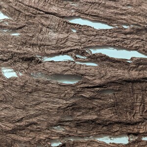 May include: Close-up of torn brown fabric strips with a textured, distressed appearance. The fabric has several horizontal openings, revealing a white background. The material appears to be thin and lightweight, with a slightly wrinkled texture.