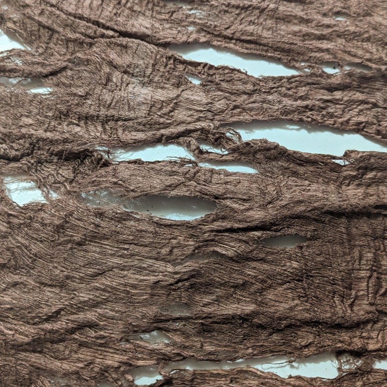 May include: Close-up of torn brown fabric strips with a textured, distressed appearance. The fabric has several horizontal openings, revealing a white background. The material appears to be thin and lightweight, with a slightly wrinkled texture.