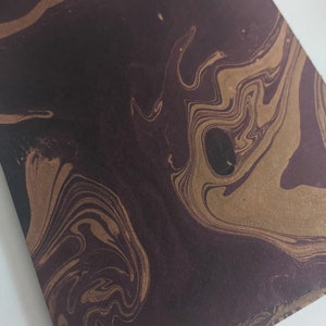 May include: Close-up of a dark maroon and gold marbled paper. The swirling pattern features shades of brown and gold, creating a unique and artistic design. The paper has a smooth texture and is ideal for stationery or craft projects.