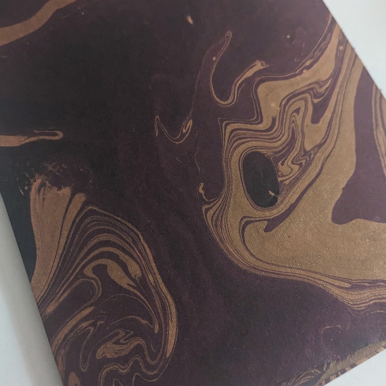 May include: Close-up of a dark maroon and gold marbled paper. The swirling pattern features shades of brown and gold, creating a unique and artistic design. The paper has a smooth texture and is ideal for stationery or craft projects.