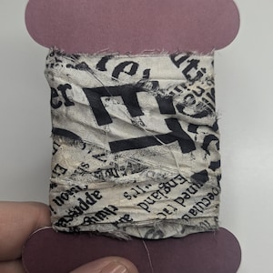 以下が含まれることがあります： 新聞のデザインのファブリックリボン。リボンは紫色のスプールに巻かれています。生地はオフホワイトで、黒い文字とぼろぼろのほつれたエッジがあります。「England」と「peculiar」という単語が含まれています。