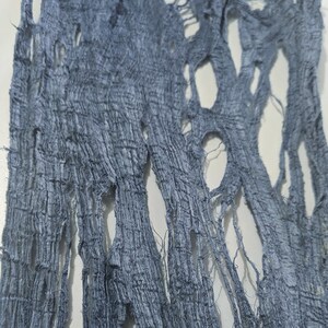 May include: Close-up of a piece of blue fabric with a distressed, shredded design. The fabric has a textured, woven appearance with numerous irregular holes and frayed edges, creating a unique, artistic aesthetic.
