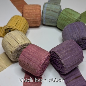 Könnte beinhalten: Eine Auswahl an Khadi-Webbandrollen in verschiedenen Farben, darunter Gelb, Orange, Grün, Lila und Rosa. Der Text "Khadi loom ribbon" ist sichtbar. Das Band besteht aus einem strukturierten Stoff, ideal für Bastelarbeiten.