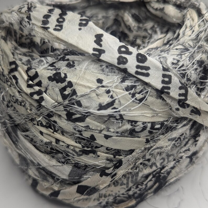 Peut inclure: Une boule de bandes de tissu en cr&egrave;me, gris et noir. Le tissu est imprim&eacute; de texte et de motifs abstraits. La texture est rugueuse et superpos&eacute;e, avec des bords effiloch&eacute;s. L'ensemble donne un textile fait main.