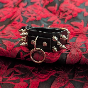 May include: Black leather choker with silver-colored cone studs and a metal ring. The choker has a buckle closure and is set against a red and black floral fabric background. A bold accessory.