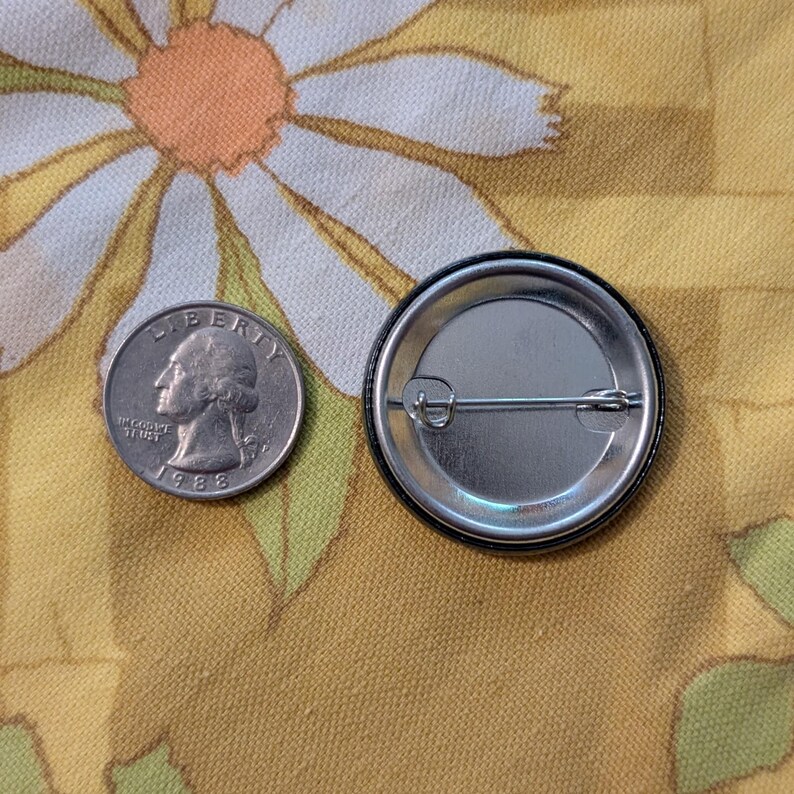 May include: A silver quarter coin next to a black and silver button. The button has a pin back. The quarter is dated 1988. The background is a yellow fabric with a daisy pattern.