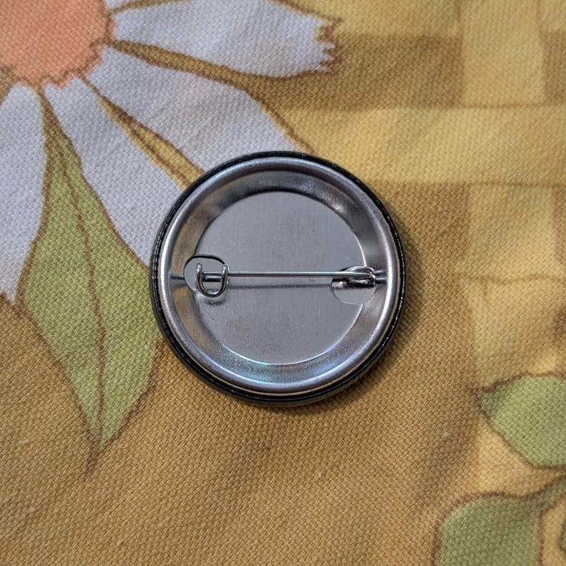 May include: A round, silver-colored pinback button with a safety pin closure. The button has a black rim and a shiny, metallic surface. The pin is designed to be attached to clothing or accessories.