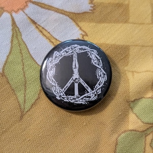 May include: A black button with a white peace sign design, encircled by a thorny wreath. The button is set against a floral patterned background with shades of yellow, orange, and green.